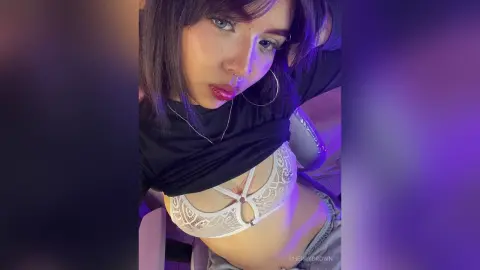 Young woman in a black top and white lace bra posing under moody purple lighting.