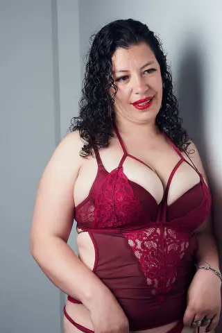 Smiling woman with curly hair wearing a burgundy lace bodysuit in a minimalist indoor setting.