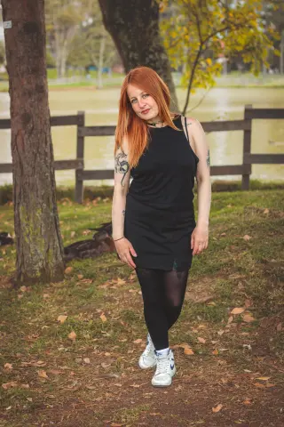 Redheaded woman in a black tank top and tights standing in a leafy autumn park by a lake.