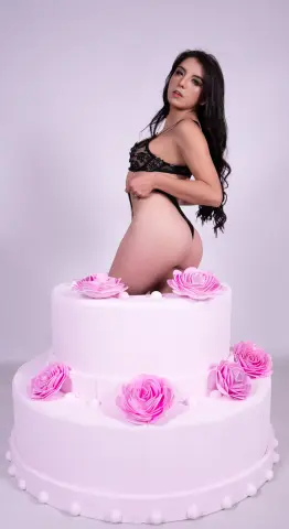 Young woman in black lace lingerie posing gracefully behind a large pink rose decorative cake.