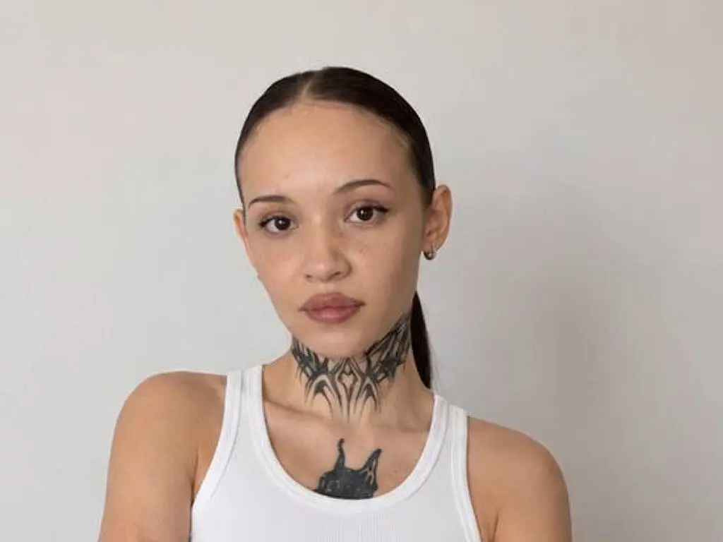 Young woman with neck tattoos wearing a white ribbed tank top against a plain neutral background.