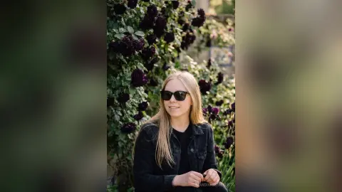 Smiling blonde woman in a black denim jacket wearing sunglasses in a lush garden with dark roses.