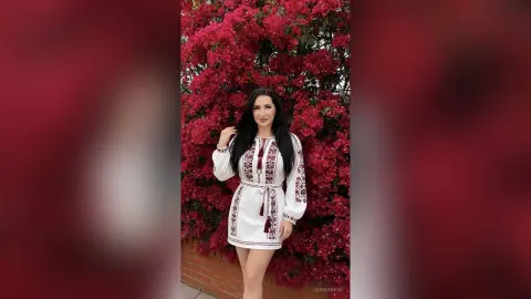 JasminNise poses in front of bright red flowers wearing a white long-sleeved dress with red embroidery.