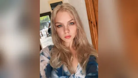 Blonde woman in a blue tie-dye blouse posing with a soft, natural look in a bright indoor setting.