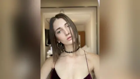 Young woman with long brown hair wearing a black lace choker and dark camisole in a bright indoor setting.