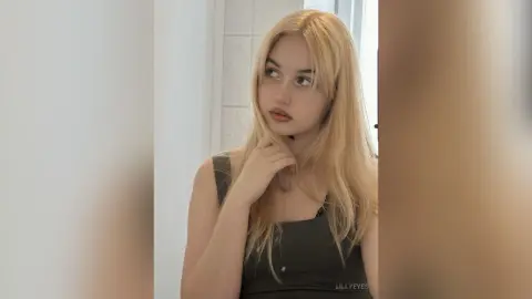 LillyEyes poses with her hand under her chin, wearing a black tank top and showing her long blonde hair.