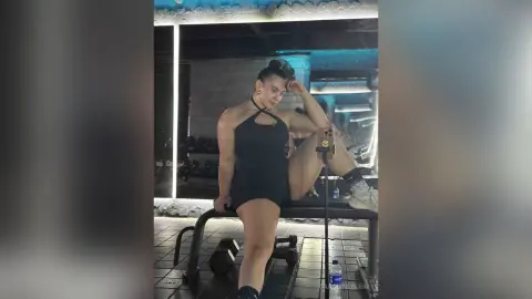 Athletic woman in black halter bodysuit posing confidently on a gym weight bench during a workout session.
