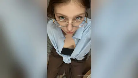 Young woman with glasses wearing a light blue button-down shirt and brown trousers in a bright indoor setting.