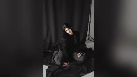 Moody portrait of a woman in a black outfit sitting on draped fabric against a dark studio background.