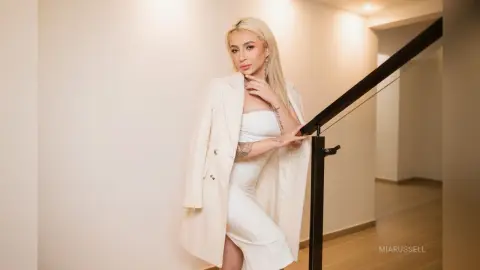 MiaRussell is a blonde woman wearing a white dress and a white blazer, posing by a black railing in a bright room.