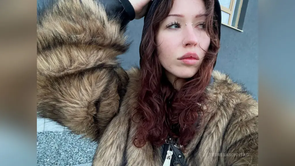 TerresaSonntag — Young woman with red hair wearing a cozy brown faux fur coat and black hoodie in an urban outdoor setting.