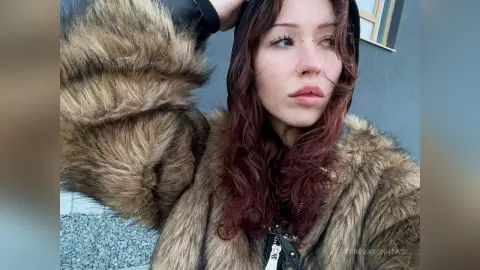 Young woman with red hair wearing a cozy brown faux fur coat and black hoodie in an urban outdoor setting.
