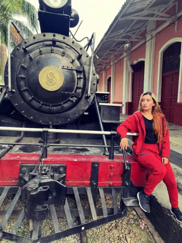 Ninablaze27 sits on the red bumper of a large black steam locomotive, wearing a red tracksuit and black top.