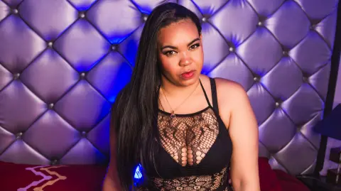 Young woman in a black lace bodysuit sitting against a tufted purple headboard in moody blue lighting.