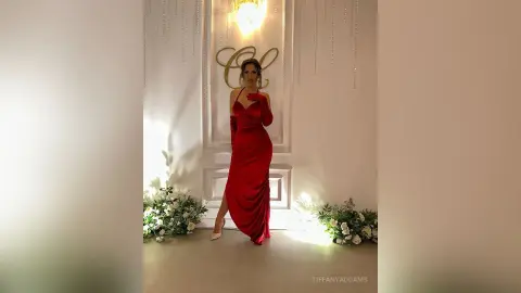 TiffanyAddams poses in a long, tight red dress with a high slit, standing in front of a white door and gold decor.