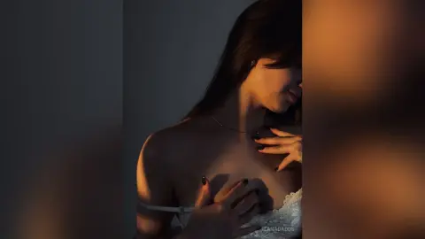 TanaDadds poses in soft lighting, wearing white lace lingerie while looking down with her hands resting on her chest.