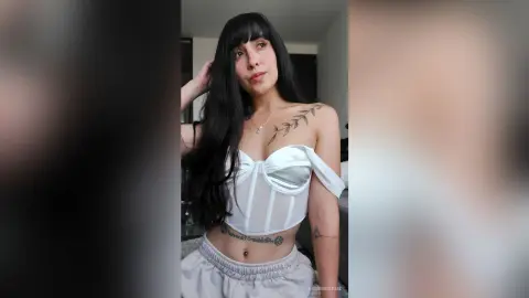 RossiNoleen poses in a white corset top and light grey lounge pants, showing off her long black hair and tattoos.