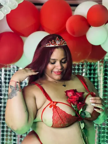 Smiling woman in red lace lingerie holding red roses against a festive red and white balloon background.
