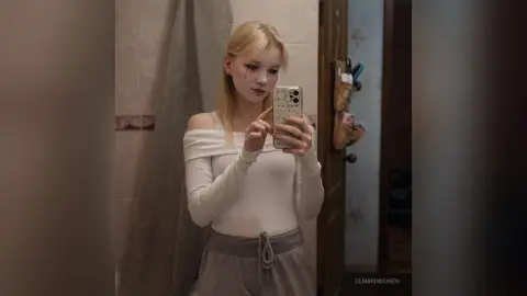 Blonde woman in a white long-sleeve top taking a moody mirror selfie in a dimly lit bathroom.