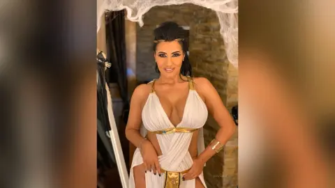 MissyJolie poses in a white and gold toga-style outfit with a deep cleavage, standing against a textured stone wall.