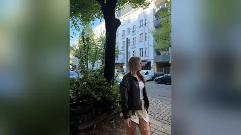 Stylish woman in a black leather jacket and white top standing on a sunny city sidewalk near green trees.