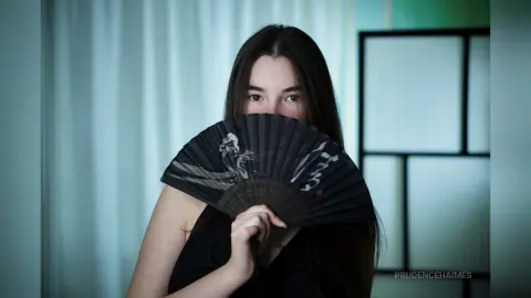 Mysterious young woman in a black dress holding a decorative folding fan in a soft-lit indoor setting.