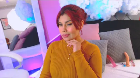 Queenamazon poses in a mustard yellow ribbed sweater against a soft pink and blue lit bedroom background.