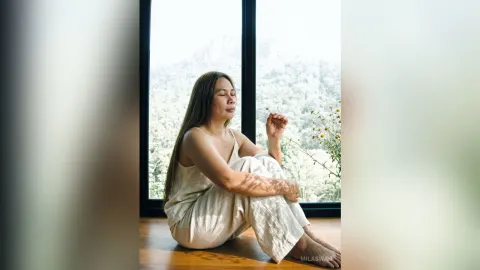 MilaSwan — MilaSwan sits on a wooden floor in white loungewear by a large window overlooking a forest.