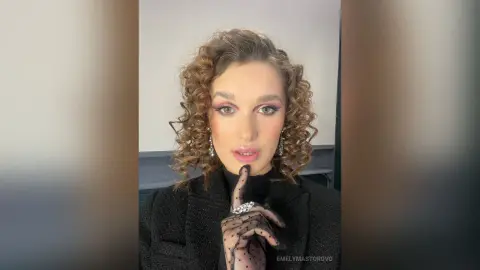 EmelyMastorovo poses with her hand near her lips, wearing a black turtleneck and sheer black gloves with curly brown hair.