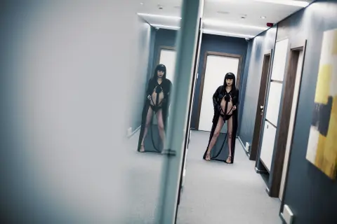 Elegant woman in a black lace bodysuit and sheer cape standing in a modern minimalist hallway.