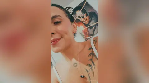 RossiNoleen smiles while looking off-camera, showing her face, dark hair, and a tattoo on her shoulder and chest.