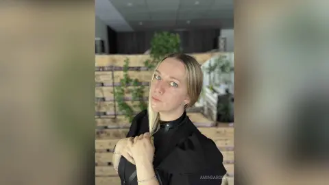 Blonde woman in a black blouse posing with a thoughtful expression in a modern indoor setting.