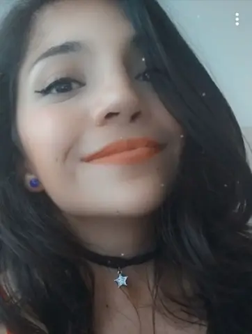 Young woman with long dark hair and orange lipstick wearing a black star choker in a soft indoor setting.