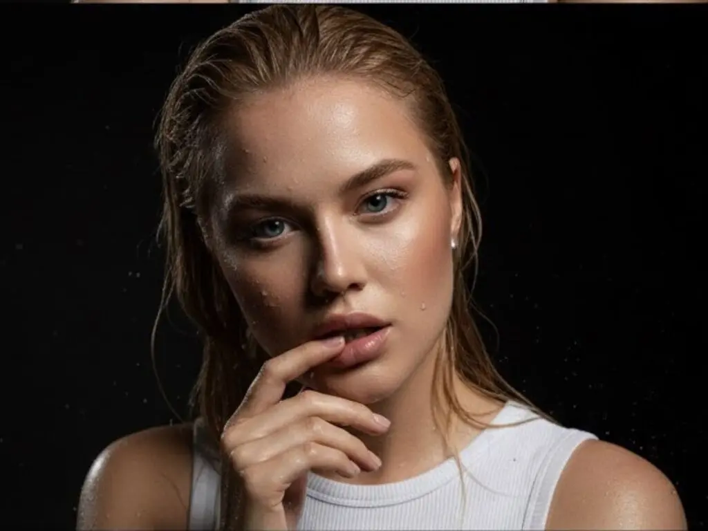 Close-up portrait of a blonde woman with wet hair and glowing skin wearing a white ribbed tank top against black.