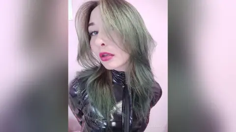 Young woman with long green hair wearing a black shiny latex top in a soft pink room.