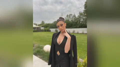 Elegant woman in a black cutout jumpsuit and blazer posing outdoors in a lush green garden setting.