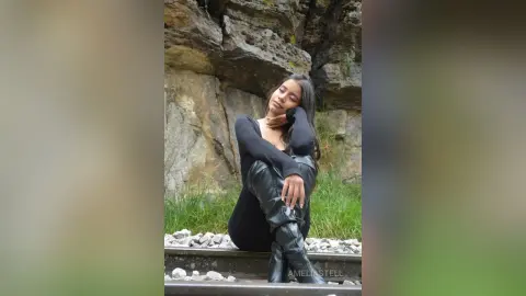 AmeliaStell poses outdoors against a rocky cliffside wearing a black long-sleeve bodysuit and black leather boots.