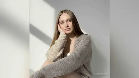 AuroraCruise poses with long straight hair, wearing a light tan knit sweater and a simple necklace against a white wall.