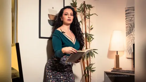 Woman in a teal top and floral skirt reading a book in a cozy, sunlit room.