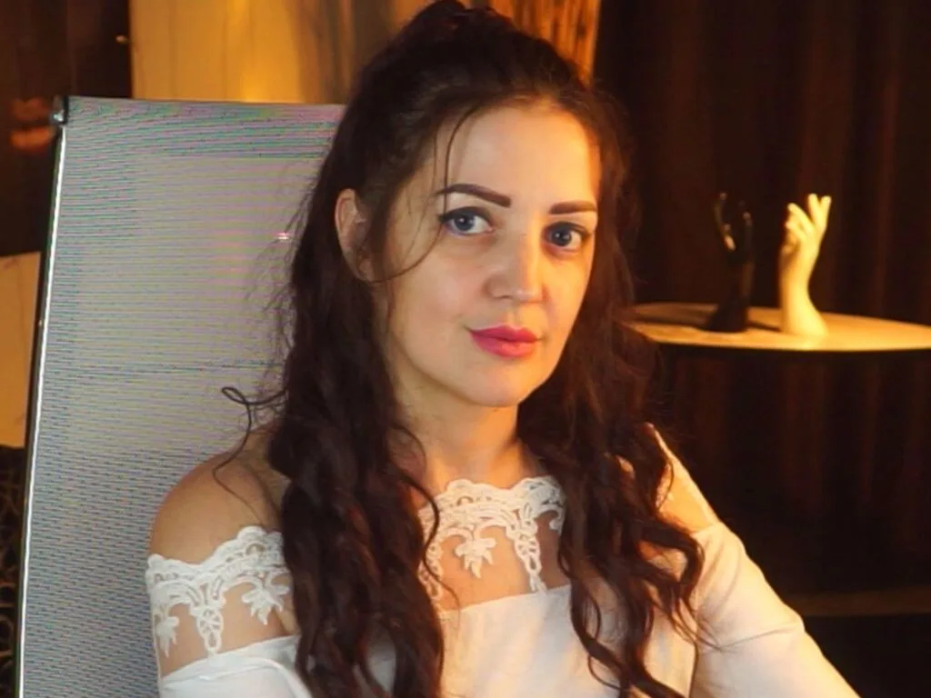 AureliaVanes — AureliaVanes poses in a white lace off-the-shoulder top with her long brown hair styled in loose waves.