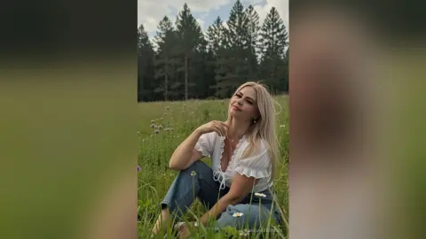 Smiling blonde woman in a white ruffled blouse sitting in a dreamy wildflower meadow by a pine forest.