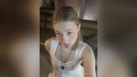 EvelinaMoorina sticks her tongue out while wearing a white tank top and a necklace in her living room.