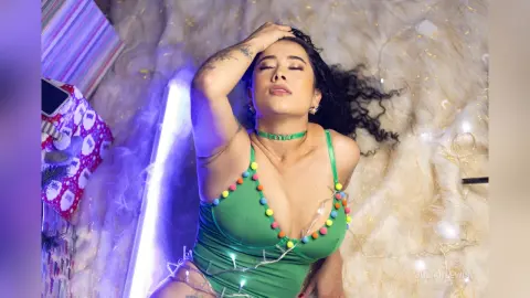 JuliahLewis lies back with her eyes closed, wearing a green low-cut bodysuit with colorful pom-poms and a matching choker.
