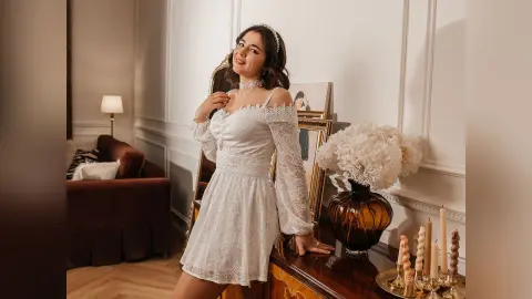 Smiling woman in a white lace cocktail dress posing in an elegant, warm-lit living room.