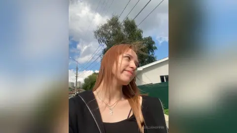 Smiling woman with long red hair in a black zip-up hoodie outdoors under a cloudy sky.