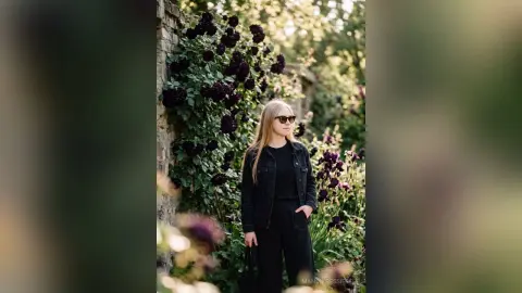 Blonde woman in a black denim jacket and sunglasses standing by a lush garden with dark purple flowers.