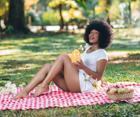 Smiling Black woman in a white dress enjoying a sunny picnic in a lush green park with a fruit basket.