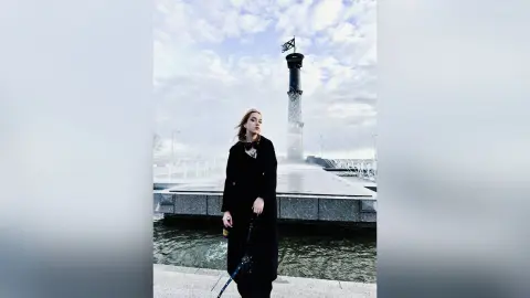OksiKoxy stands outdoors in a black coat and sunglasses near a waterfront monument under a cloudy sky.
