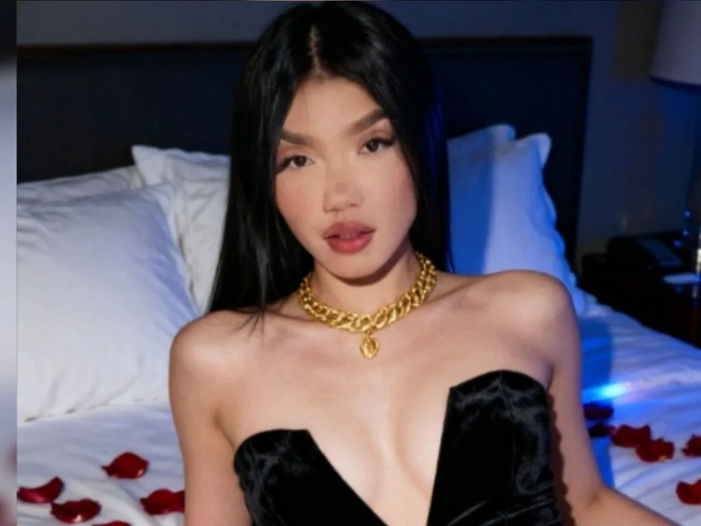 AnastasiaVelo — AnastasiaVelo wears a black velvet dress and a gold chain necklace while sitting on a bed with white pillows and red rose petals.