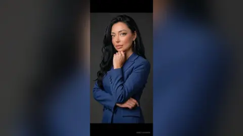AnneThompson poses with her hand near her chin, wearing a blue blazer and hoop earrings against a dark background.